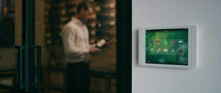 A home automation touchpad sits on a wall. In the background, a man chooses a wine bottle from a wine room.